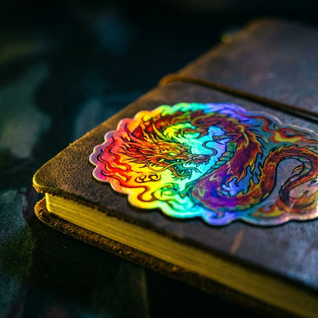 Manos sosteniendo un sticker holográfico bajo luz amarilla, un arcoíris inunda el material.